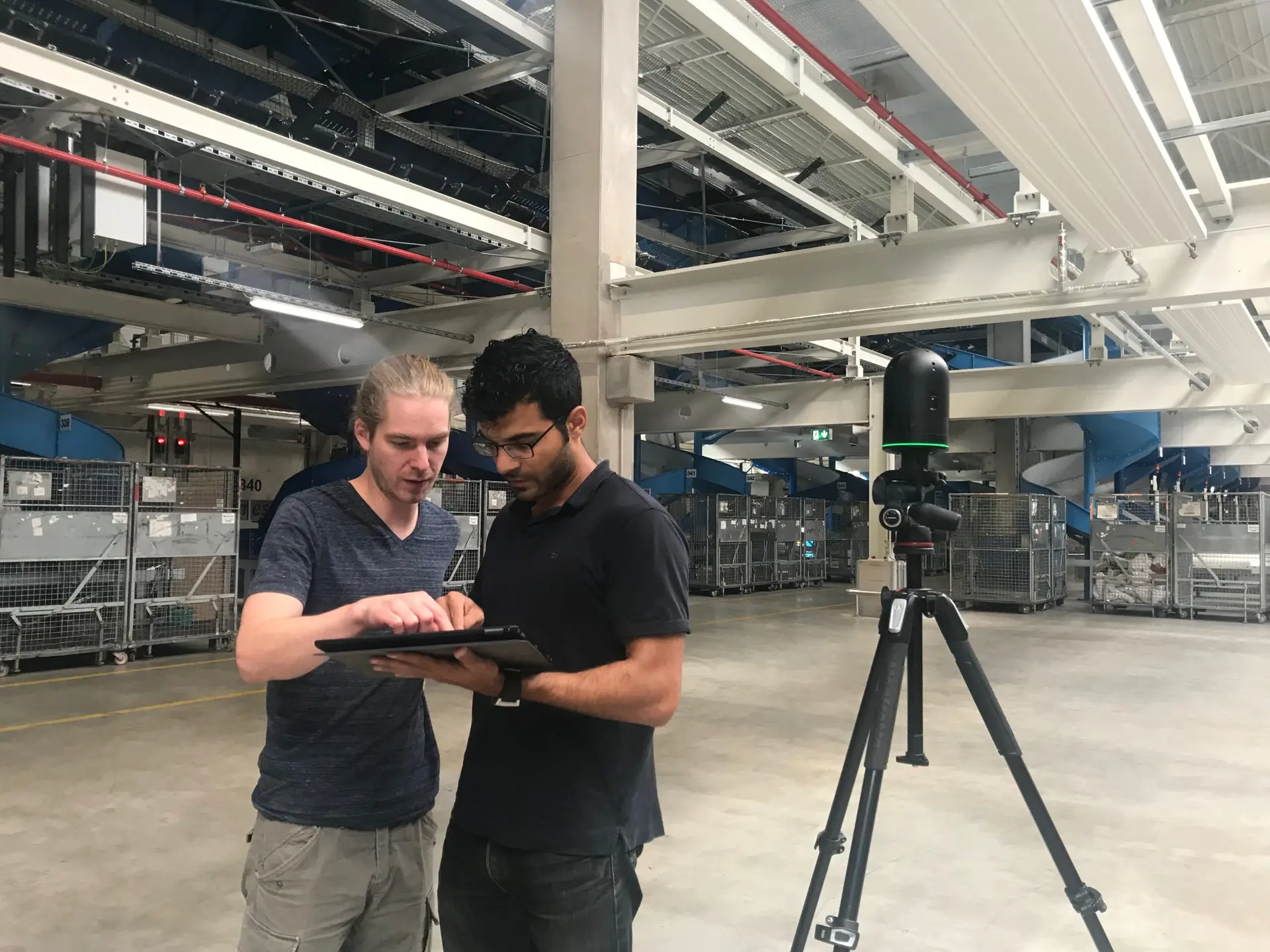 Zwei omnia360-Teammitglieder prüfen am Tablet die Aufnahme, daneben ein 3D-Scanner auf Stativ in einer Logistikhalle